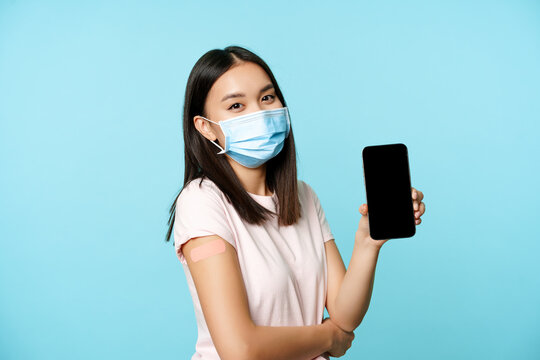 Asian woman in medical face mask, showing arm with patch after covid-19 vaccine, smartphone screen with app interface, concept of vaccination certificate, health passport