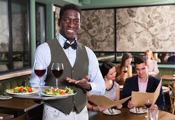 Hospitable African American cheerful smiling waiter standing with serving tray, recommending dishes in restaurant