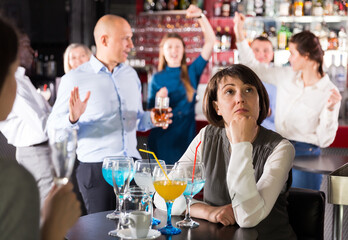 Sad woman sitting alone at office party in nightclub in background with cheerful workmates
