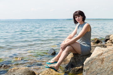 brunette woman in a striped summer dress sits on rock against the background of the sea.