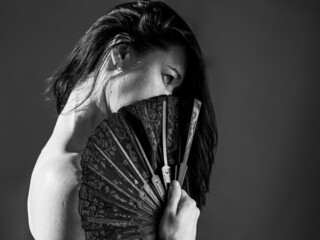 Portrait of beautiful attractive half face brunette woman with fan covering her face, bare shoulders. Black and white photo on black background. - obrazy, fototapety, plakaty