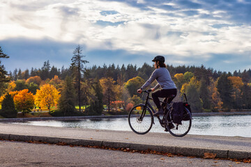 White Caucasian Adult Woman riding a bicycle on Seawall in Stanley Park. Cloudy Fall Season. Downtown Vancouver, British Columbia, Canada.