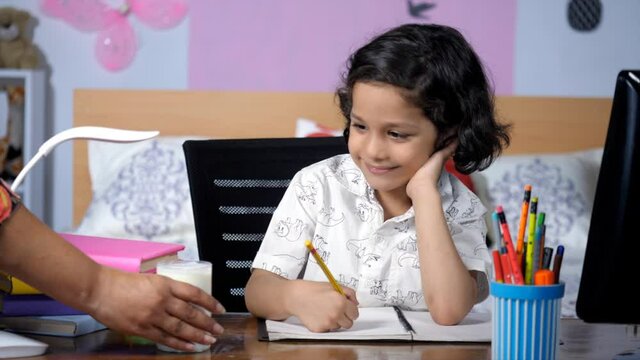 A loving affectionate mother offering a glass of milk to her little son in the morning. Closeup shot of a sweet kid in casual clothes doing his homework / studying sitting on his desk at home