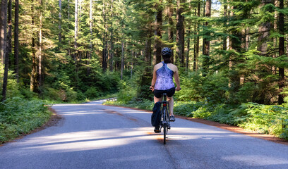 Adventurous White Caucasian Woman on a bicycle riding on a path in Green Canadian Rain Forest. Seymour Valley Trailway in North Vancouver, British Columbia, Canada.