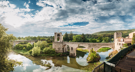 Besalu, Girona, Catalonia, Spain. Famous Landmark Old Medieval Romanesque Besalu Bridge Over The Fluvia River In Cloudy Summer Day