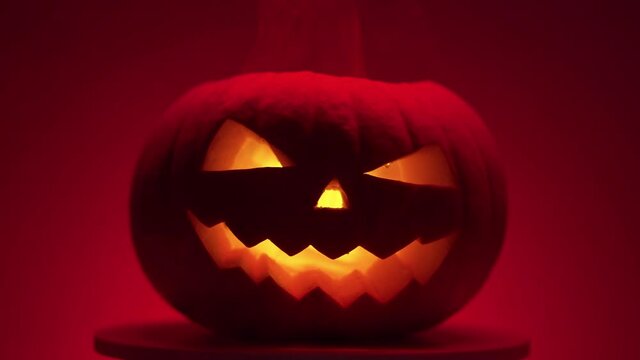 halloween orange smiling glowing smoking pumpkin with carved teeth, eyes and nose on a round light platform in red light on a dark background