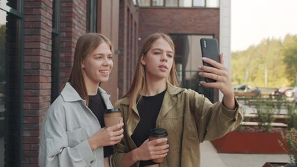 Tracking shot of cheerful young twin sisters holding coffee in to-go cups and recording vlog on their mobile phone outside - Powered by Adobe