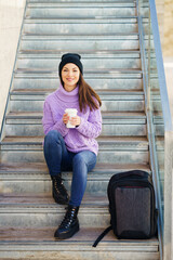 Woman in her twenties taking a coffee break sitting on some steps in the street.