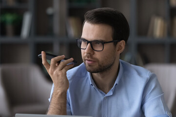 Concentrated young businessman in eyewear recording audio message on cellphone, sharing ideas in...