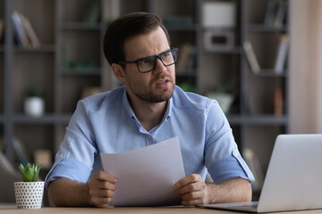 Stressed young businessman in eyewear feeling nervous of getting bad news in paper correspondence, reading letter with bank loan rejection or bankruptcy notification, having financial problems.