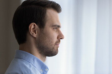 Obraz premium Close up head shot side view dreamy confident young handsome man looking in distance out of window, visualizing successful future, planning weekend or vacation trip, meditating alone at home.