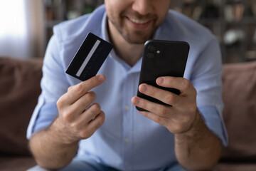Close up happy young man holding credit card and phone in hands, entering payment information in mobile shopping app, purchasing goods services online, satisfied with successful financial operation.