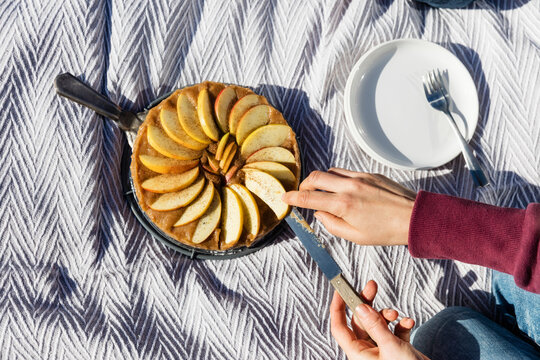Hands of young woman cutting freshly baked apple pie on picnic blanket