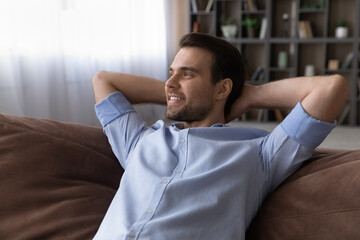 Obraz premium Relaxed young man lying on comfortable sofa with folded hands behind head, looking in distance daydreaming visualizing future, enjoying peaceful carefree leisure weekend moment alone in own home.