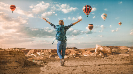 Young Handsome Tourist Mountain Hiking with Rucksack. Male Adventurer Raises His Hands Up and Celebrates Nature on Top of a Rocky Canyon Valley. Hot Air Balloon Festival in the Sky of National Park.
