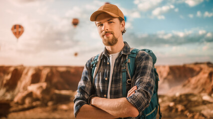 Fototapeta premium Portrait of a Young Handsome Tourist Posing with Backpack on Top of a Rocky Canyon Valley. Adventurous Male Backpacker on a Hiking Journey Trip. Hot Air Balloon Festival in Mountain National Park.