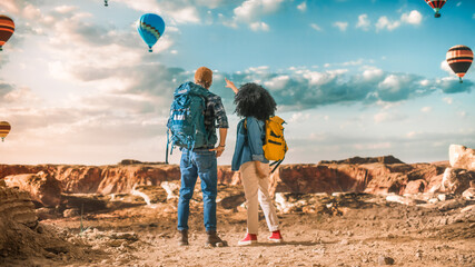 Young Diverse Tourist Couple Hiking with Backpacks in Great Wilderness in Rocky Canyon Valley. Male and Female Backpacker Friends on Adventure Trip. Hot Air Balloon Festival in Mountain National Park.