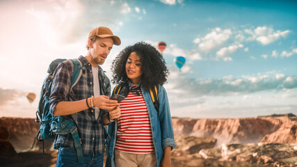 Young Diverse Couple of Hikers are Checking Tourist Information and Directions on Their Smartphone on Top of Rocky Canyon Valley. Backpackers on Adventure. Hot Air Balloon Festival in National Park.
