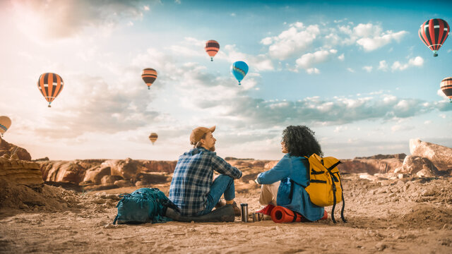 Young Diverse Tourist Couple Hiking with Backpacks, Resting on Top of Rocky Canyon Valley. Male and Female Backpacker Friends on Adventure Trip. Hot Air Balloon Festival in Mountain National Park.