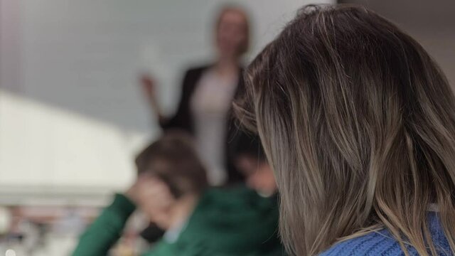 Back view of a woman in a lecture hall, listening to a professor giving a class with whiteboard at the background.