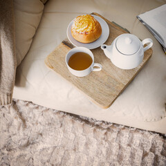 Teapot, cup of tea  and bun on the wooden try, open book on cozy sofa for relax time at home.