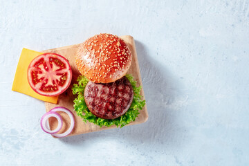 Beef burger ingredients on a wooden board with a place for text, shot from above. Grilled patty, lettuce leaf, tomato, Cheddar cheese, onion and a sesame bun