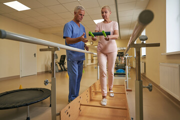 Physiotherapist helping mature patient walk between parallel bars in hospital