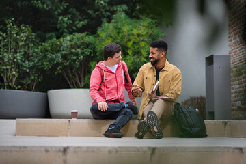 Happy young man with Down syndrome and mentoring friend sitting and talking outdoors