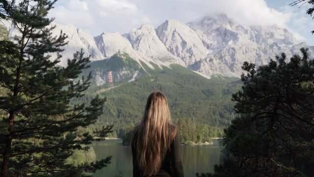 A blonde girl with long hair is standing in front of lake Eibsee in Germany. She is watching the beautiful backdrop. There are mountains with snow o top of them.