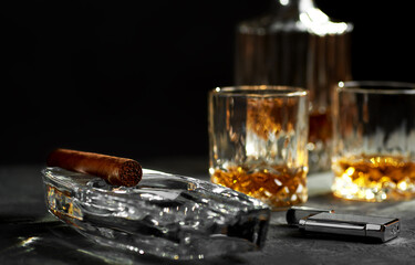 Still life with a Cuban cigar in an ashtray, a glass and a bottle of whiskey and a lighter on a stone table top.