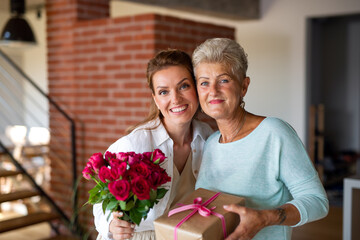 Happy senior mother getting gift and bouquet from adult daughter indoors at home, looking at camera.
