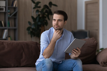 Thoughtful young handsome man looking in distance holding digital tablet in hands, web surfing information, spending free leisure time online, sitting on comfortable sofa in modern living room.