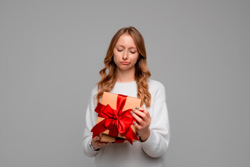 Young woman in white sweater opening birthday gift on party celebration, looking disappointed unhappy, standing over gray background. Holidays, celebration and lifestyle concept
