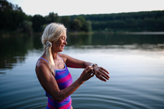 Portrait of active senior woman swimmer standing and setting smartwatch outdoors in lake.