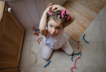 High angle view of small girl applying hair curlers indoors at home, looking at camera.
