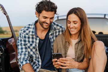 White couple smiling and using cellphones while sitting in car trunk