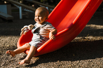 Fototapeta na wymiar White toddler wearing overalls sitting on slide with toy at playground