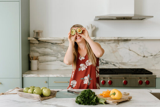 Happy female nutritionist covering eyes with avocado in kitchen