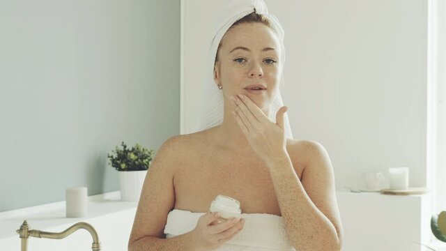 Portrait of  woman wrapped in towel in bathroom. female, after waking up and taking shower, takes moisturizing cream from jar and is applied to  face and rubs it into skin to give softness.