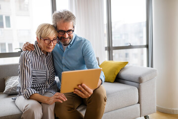 Beautiful happy mature couple is using a digital tablet, talking and smiling at home