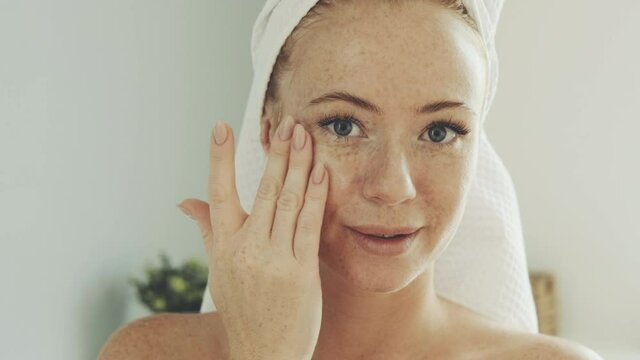 Close-up of face of young woman with a towel on her head in bright white bathroom. female takes moisturizing cream from jar and is applied to face and rubs it into skin to give softness.