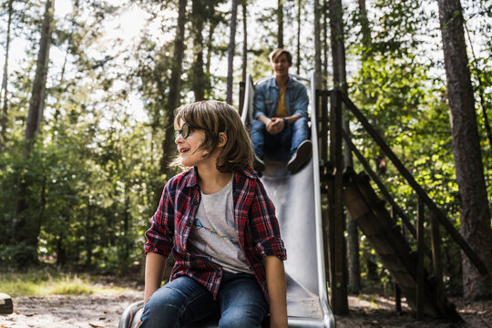 Smiling son sitting on slide with father in background at forest