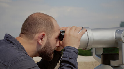 Fototapeta na wymiar Close up of caucasian man looking through telescope lens at panoramic view of city from observation point on building roof. Young person using binoculars for outdoor panorama sightseeing