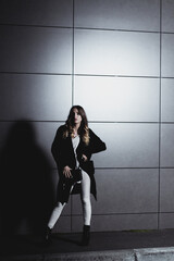 Sexy girl posing against a dark wall in the evening. An elegant fashionable girl in a long black coat, white jeans, with a leather bag stands near the wall of the building. Beautiful girl for a walk