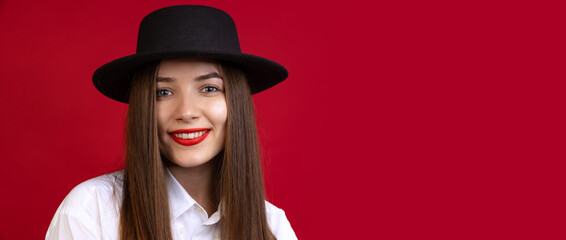 Close-up of stylish young beautiful girl wearing black hat isolated on dark studio background. Concept of emotions, fashion