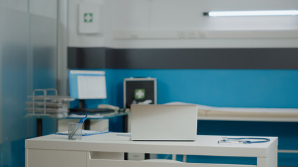 Empty medical cabinet at healthcare facility with laptop, stethoscope and checkup papers on white desk. Nobody in professional interior for consultation with modern furniture and tools