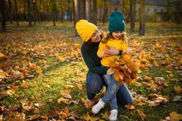 Happy motherhood, carefree childhood. Cheerful loving mother hugs her lovely cute baby girl sitting on her knees while relaxing in the autumn park and gathering a bouquet of falling dry maple leaves