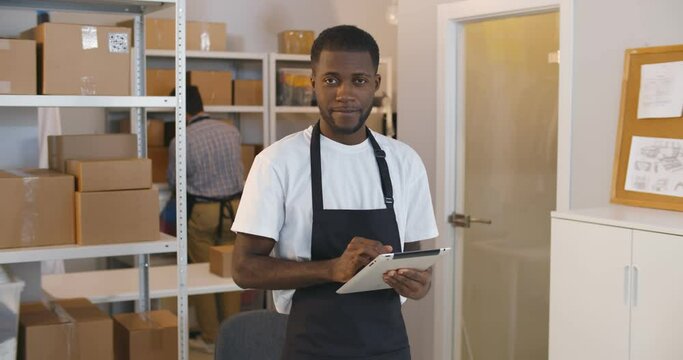 Portrait of young mixed-race man using digital tablet in warehouse doing inventory smiling at camera