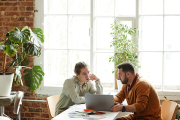 Wide angle portrait of two creative people using laptop while discussing design project in studio,...