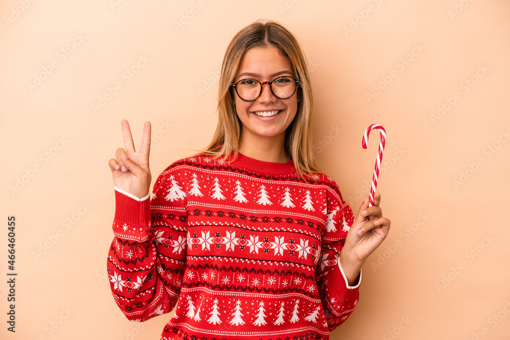 Wall mural young caucasian woman holding a christmas stick isolated on beige background showing number two with